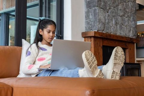 Indian girl studying on laptop reclining on tan leather sofa in cozy modern living room