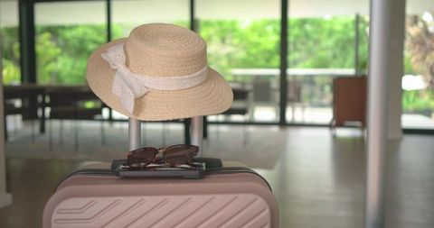 Sun hat and sunglasses resting on pink hard-shell suitcase in modern sunlit hotel lobby