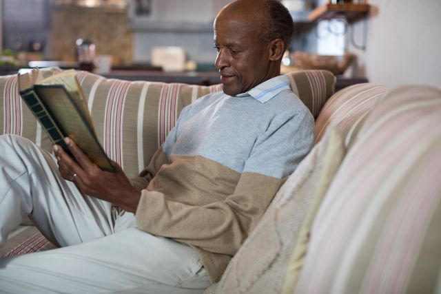 Senior man enjoying a quiet moment reading a book while sitting on a comfortable sofa in his living room. Ideal for use in articles or advertisements related to senior living, retirement, leisure activities, and home comfort.