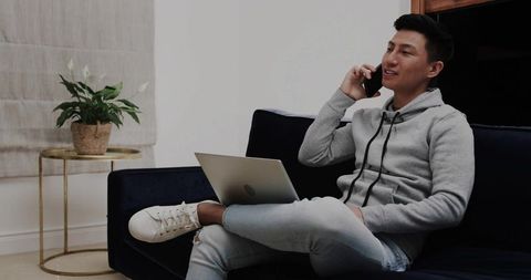 Asian man working from home on laptop while talking on smartphone on sofa