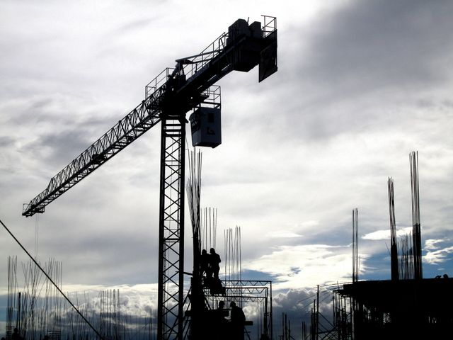 Silhouetted view of a construction site featuring a tall tower crane against a dramatic cloudy sky. Several steel beams stand vertically, creating a scene of active building and infrastructure development. This image is perfect for illustrating industrial themes, architecture elements, and engineering projects in urban developments.