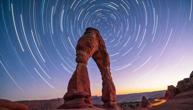 Sandstone arch framing spiral star trails over desert plateau at twilight with light trail