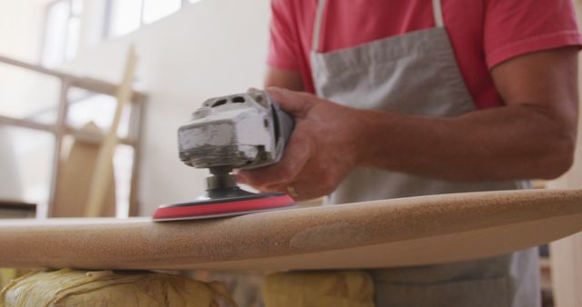 Craftsman using sanding machine on surfboard in workshop, emphasizing skill and precision. Suitable for materials involving DIY projects, craftsmanship, surfboard making, detailed manual labor. Ideal for articles or advertisements related to woodworking, artisanal creations, surf culture.