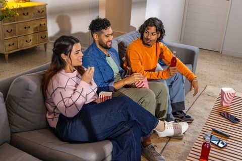 Multicultural friends watching movie on couch sharing popcorn and bottled drinks at home