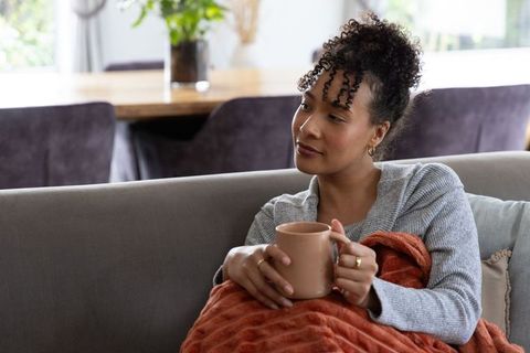 African american woman relaxing on sofa holding mug wrapped in cozy rust blanket