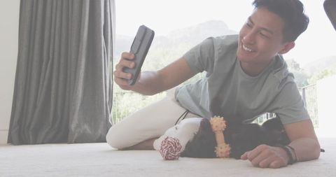 Young man smiling while taking selfie with playful cat on sunlit living room carpet
