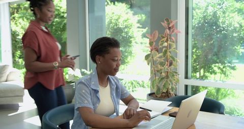 African mother and daughter working remotely at sunlit home with laptop and smartphone