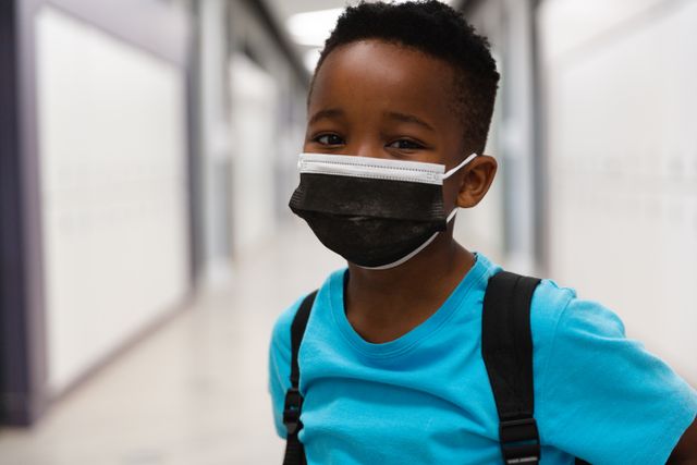 This image shows an African American elementary schoolboy wearing a mask while standing in a school corridor. It can be used for educational materials, health and safety campaigns, back-to-school promotions, and articles related to childhood education during the pandemic.