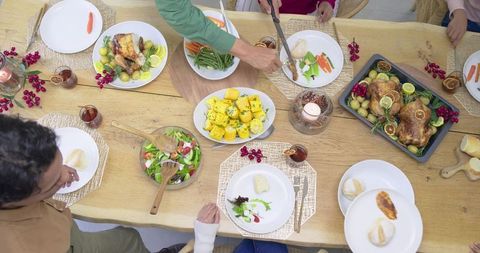 Multigenerational diverse family sharing roast dinner, passing dishes around wooden table