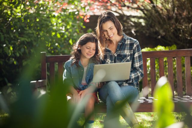 Smiling mother and daughter using laptop while sitting on wooden bench at backyard