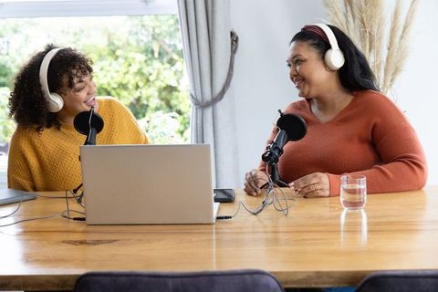 Women cohosting home podcast at sunlit table with laptop, microphones, headphones