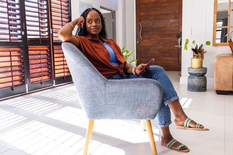 Relaxing woman sitting in bright modern living room holding phone by wooden shutters