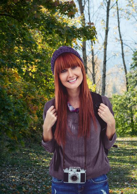 Woman traveler smiling while exploring forest with autumn colors. She wears a beanie, gray sweater, and backpack, holding vintage camera hanging around her neck. Useful for promoting travel, adventure tourism, nature exploration, travel gear, and photography equipment.
