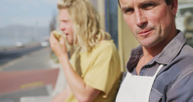 Two colleagues taking a break outdoors, with one person wearing an apron and looking into the camera, while the other enjoys a meal in the background. Ideal for use in business, food industry contexts, employee wellness programs, or casual workplace environments.