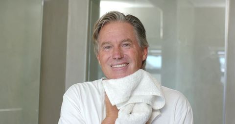 Mature man smiling while patting face with towel in bathroom mirror for morning grooming