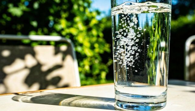Sunlit tall glass of sparkling water with rising bubbles casting crisp shadow on table