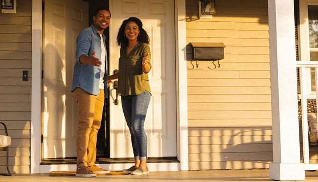 Smiling couple welcoming guests on sunlit front porch during golden hour, home entry