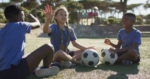 Diverse kids sitting on soccer field sharing balls and laughing during sunny team practice