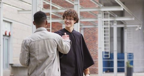Smiling graduate greeting friend in campus courtyard wearing black gown and denim jacket