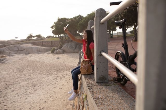 Side view of a biracial woman enjoying free time in nature on a sunny day, sitting on the wall, holding a smartphone, taking selfie, wearing smartwatch, beige backpack next to her.