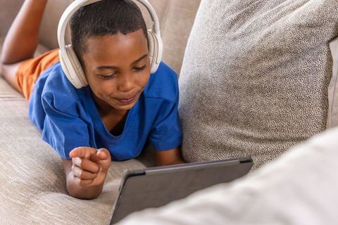 African american boy wearing headphones watching tablet while lying on cozy beige couch