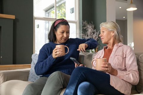 Mature diverse friends chatting on sofa with coffee, checking time on smartphone watch