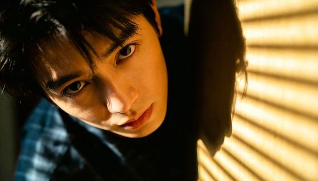 Moody sunlit cinematic portrait of young man leaning by blinds with intense gaze