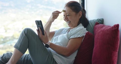 Senior Asian woman practicing facial self-care by window using jade roller and mirror