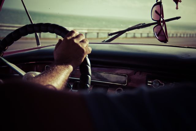 Hand gripping steering wheel of vintage car while driving along a coastal road with ocean visible through windshield, combined with hanging sunglasses hinting at sunny day. Ideal for illustrating travel, road trips, adventure, classic car restoration, or vintage styling themes.
