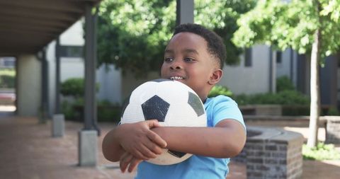 Smiling boy hugging soccer ball in sunlit courtyard, playful youth enjoying outdoor game