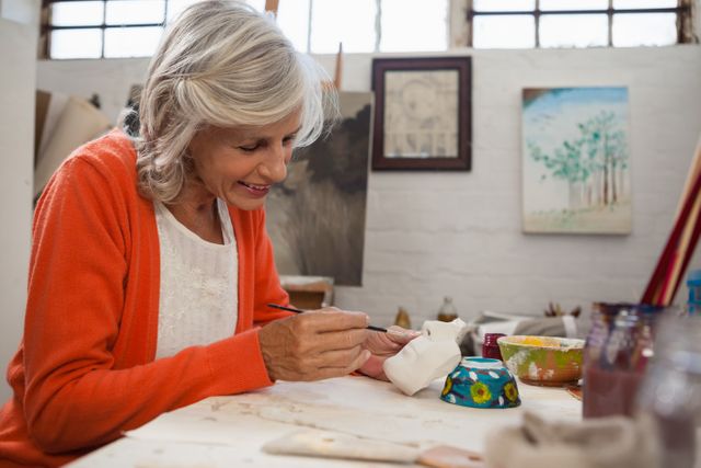 Senior woman painting a ceramic bowl in an art class. She is focused and enjoying her creative hobby. Ideal for use in articles about senior activities, creative hobbies, art classes, and promoting mental well-being through artistic expression.