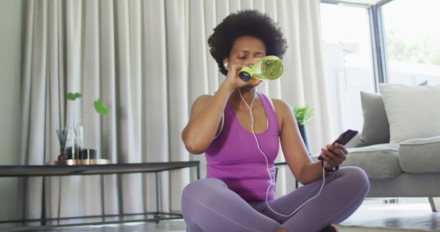 African American woman sitting on yoga mat, drinking water after home workout while holding smartphone. Ideal for promoting fitness, hydration, healthy living, and home workout routines.