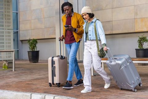 Friends walking with rolling suitcases and checking smartphone outside transit hub