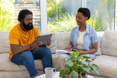African american couple planning at home using tablet and notebook on sunlit sofa