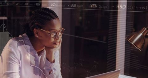 Woman analyzing financial data on laptop at night with reflective glass and currency tickers