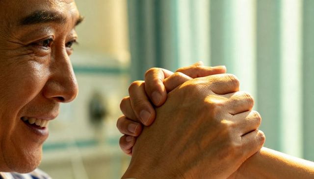 Senior asian man smiling holding interlocked hands by sunlit window conveying warmth