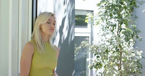 Contemplative blonde woman leaning by glass door in sunlit modern courtyard interior