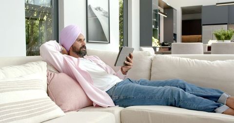 Relaxed Man in Pink Turban Browsing Tablet on Modern Sofa at Home