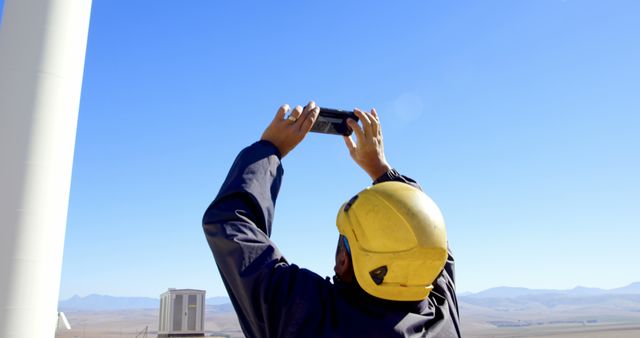 A middle-aged engineer in a hard hat and work attire is taking a photo with his smartphone, with copy space. Capturing progress or inspecting work on an industrial site, the image reflects the integration of technology in fieldwork.