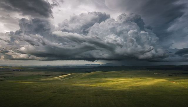Dramatic thunderstorm cloud towering over rolling prairie with sunlit meadow beams
