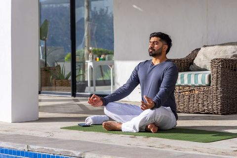 Indian man meditating on pool deck on green yoga mat during sunny outdoor wellness session