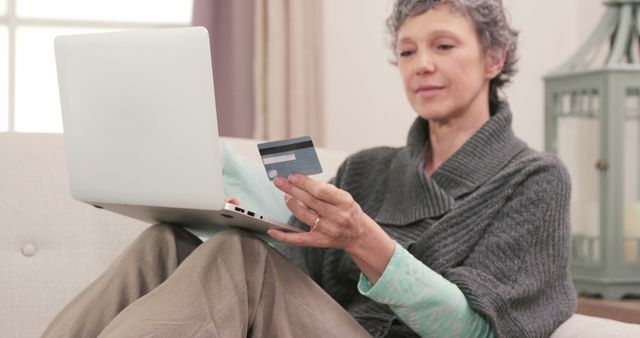A senior woman is sitting on a sofa in her living room, using a laptop to shop online with her credit card. This stock photo is perfect for illustrating concepts related to consumer technology use among older adults, e-commerce, online banking, and secure online transactions. It is ideal for use in advertisements, blogs, educational materials, and financial services marketing targeting seniors.