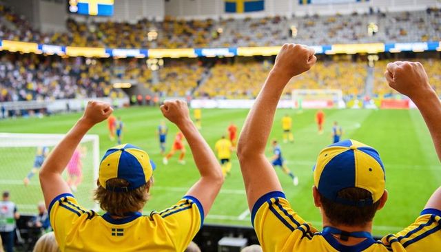 Swedish soccer fans cheering in stadium wearing yellow jerseys and blue caps, raising arms