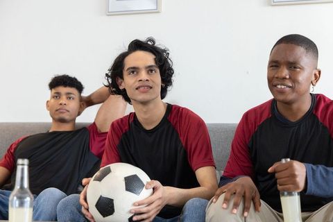 Diverse young men watching soccer at home holding soccer ball and bottled drinks