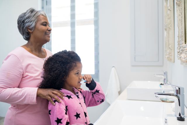 Grandmother helping granddaughter brush teeth in bathroom. Captures family bonding and daily morning routine. Ideal for use in family-oriented campaigns, health and hygiene promotions, and advertisements focusing on oral care products.