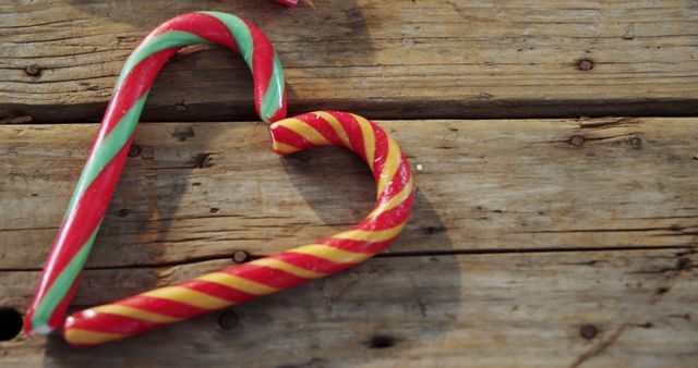 This image shows two candy canes bent together to form a heart shape on a rustic wooden surface. The vibrant red, green, and yellow candy canes evoke a festive mood, making this suitable for Christmas and holiday themed projects. This image can be used for holiday greeting cards, social media posts celebrating Christmas, or decorations for winter-themed parties and seasonal advertisements.