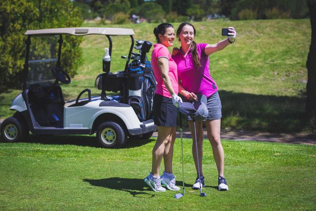 Two women are taking a selfie on a golf course on a bright sunny day. They are standing near a golf cart, holding golf clubs, and smiling. This image can be used for promoting sports, active lifestyles, outdoor activities, and friendship. It is ideal for advertisements, social media posts, and articles related to golf, leisure, and summer activities.