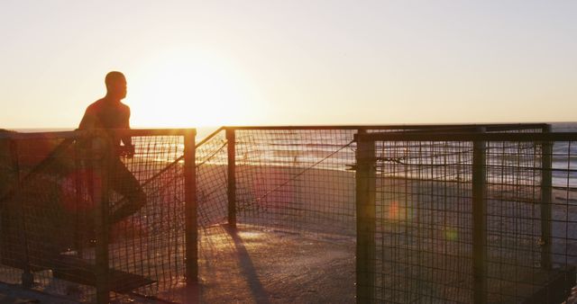 Man standing on deck overlooking ocean during sunset. Silhouette against sky, creating peaceful scene. Perfect for themes of contemplation, tranquility, nature, relaxation and solo travel.