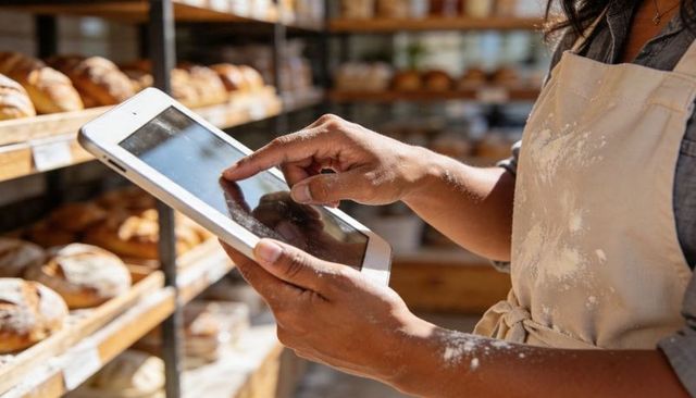 Bakery worker checking tablet while wearing flour-dusted apron beside bread shelves