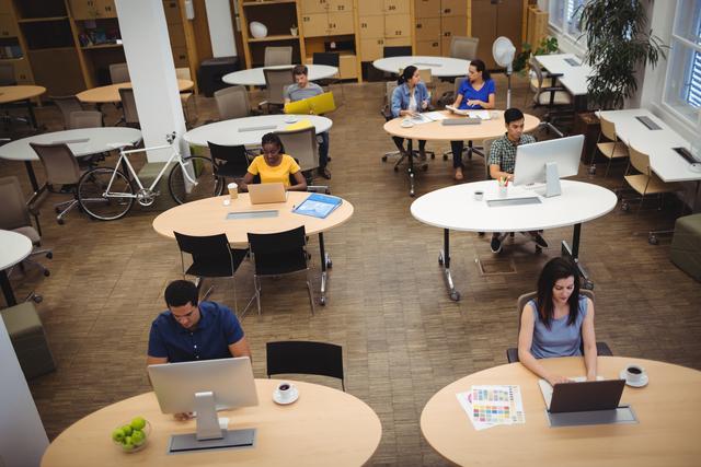 Graphic designers are working at their desks in a modern, open-plan office. The workspace features multiple desks with computers, fostering a collaborative and creative environment. This image can be used to illustrate concepts related to teamwork, productivity, modern office settings, and the creative industry.